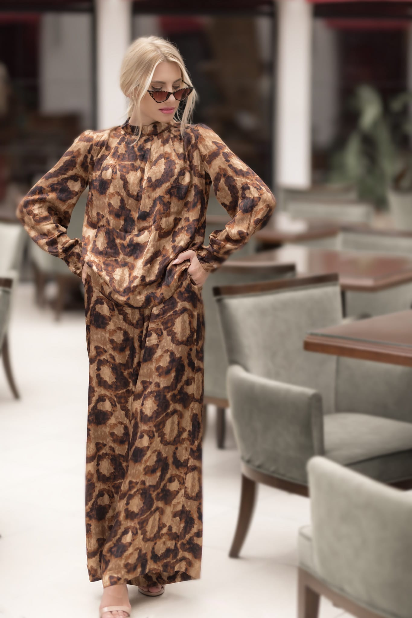 model in a leopard print dress standing in an indoor setting with chairs and tables.