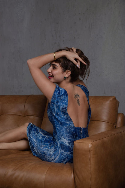 Woman in a blue dress sitting on a brown leather couch against a gray wall.