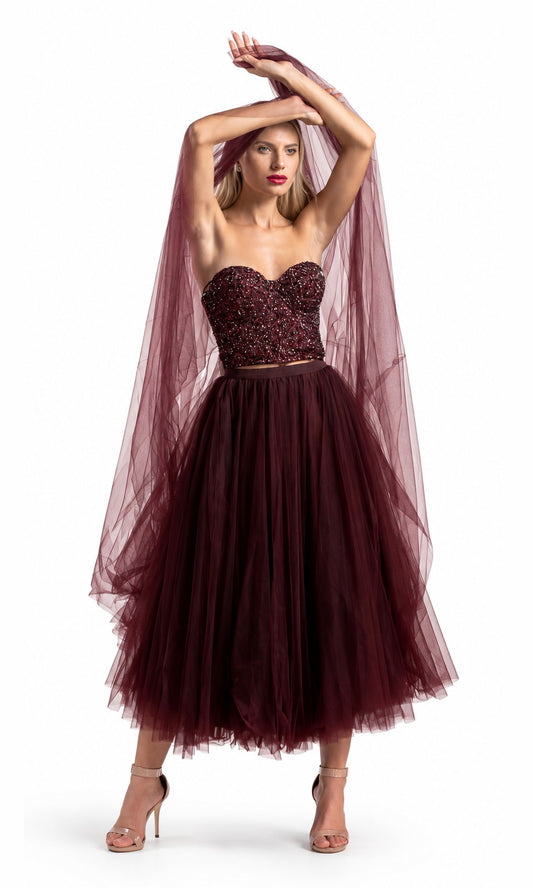 A woman modeling a strapless tulle dress in deep burgundy, featuring a heavily embellished beaded sweetheart bodice and a voluminous layered tulle skirt, with a matching sheer tulle veil draped over her head against a white studio background.