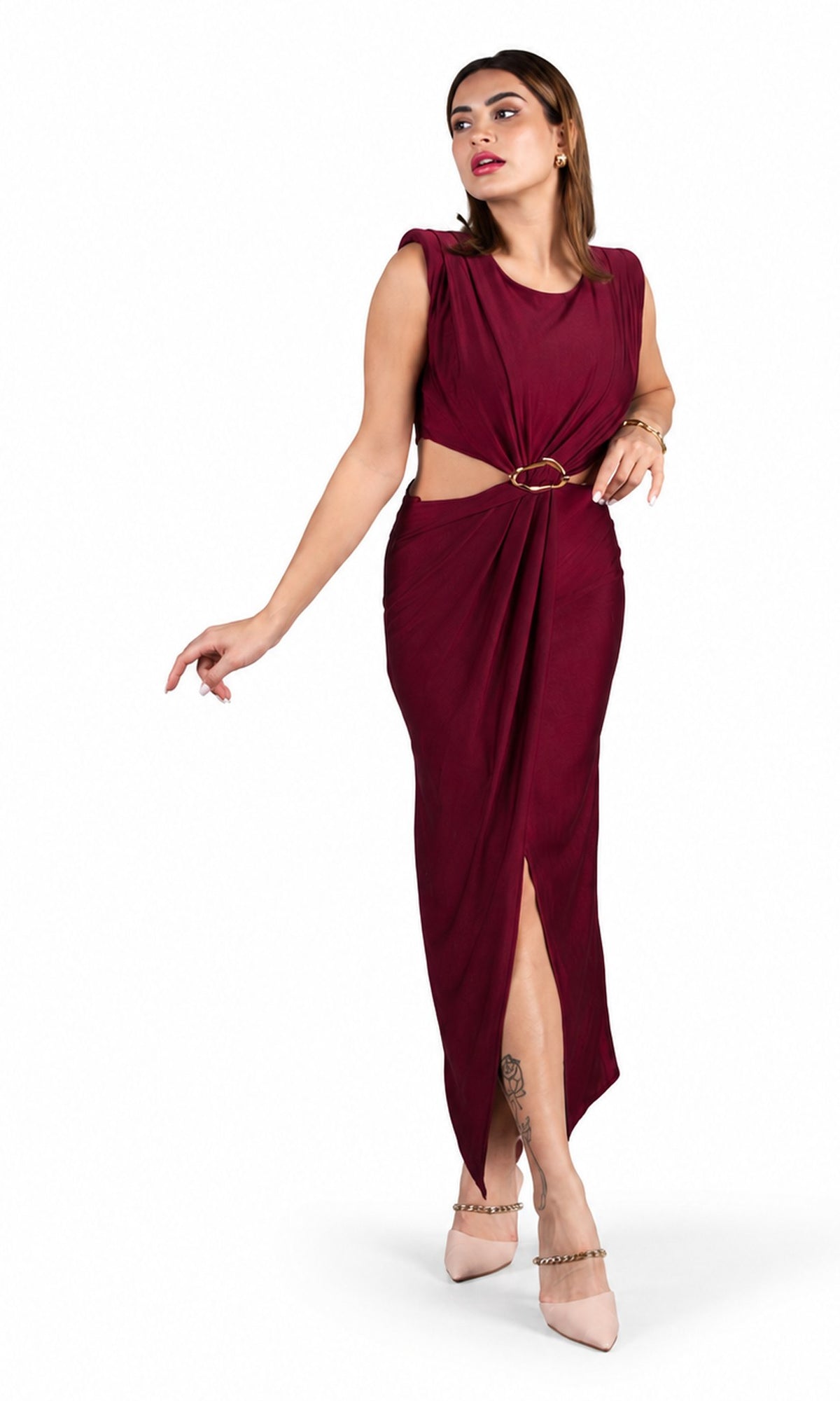 A woman models a sleeveless, deep burgundy maxi dress with a cutout at the waist, a gold ring accent, and a high side slit. She is wearing nude heels and standing against a plain white background.
