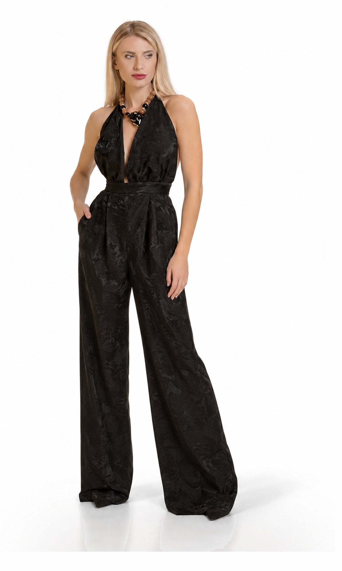 A blonde model wearing a black halter-neck jumpsuit with a subtle jacquard pattern, standing against a plain white studio background.