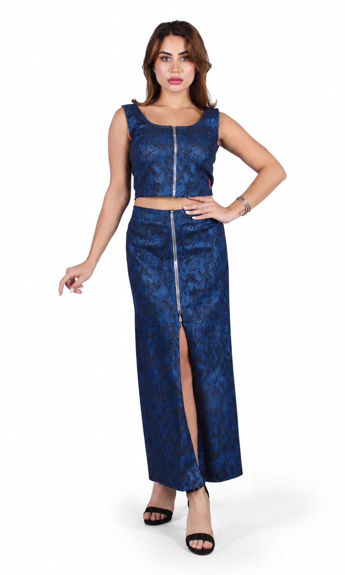 A woman modeling a two-piece blue patterned set consisting of a sleeveless crop top with a front zipper and a matching high-slit maxi skirt, photographed against a plain white studio background.