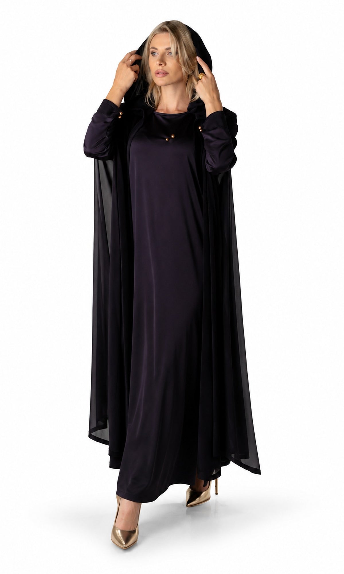A woman modeling a long, flowing black dress with matching sheer draped sleeves and a matching head covering, styled with gold high heels against a plain white studio background.