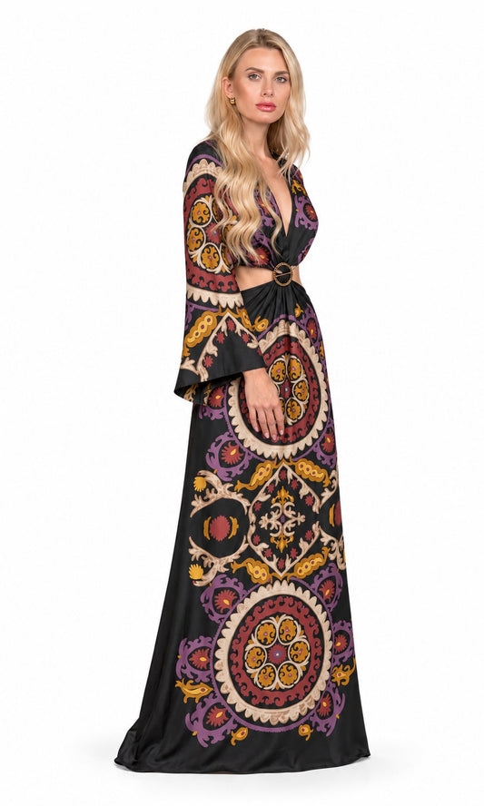 A woman modeling a long black maxi dress with a colorful bohemian mandala pattern in purple, orange, yellow, and beige. The dress features a deep V-neckline, a matching black belt with gold hardware, and flared bell sleeves.