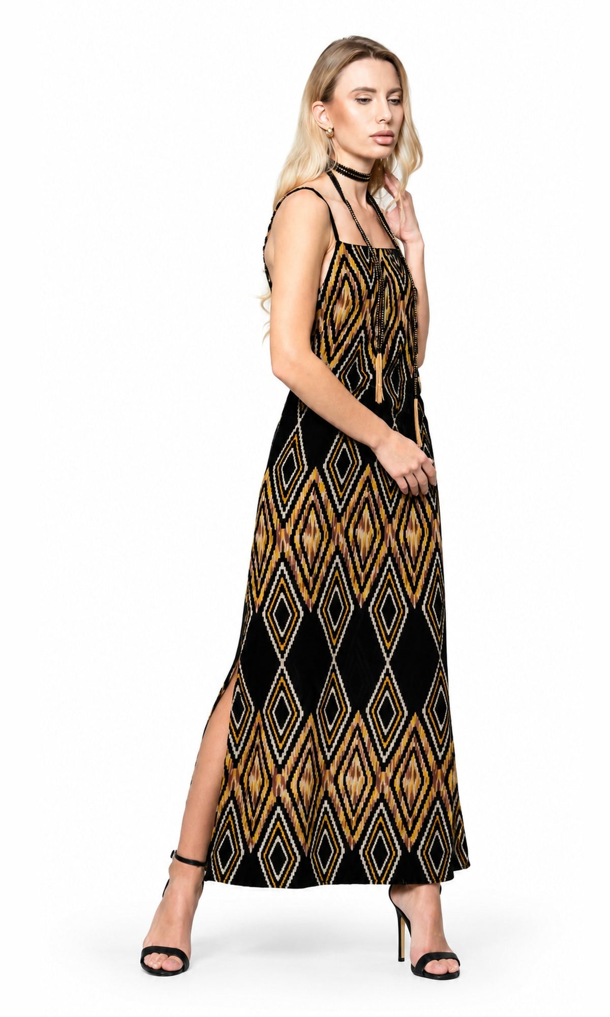 A woman modeling a sleeveless black maxi dress with a gold and beige geometric diamond pattern, styled with black strappy heels against a plain white background.