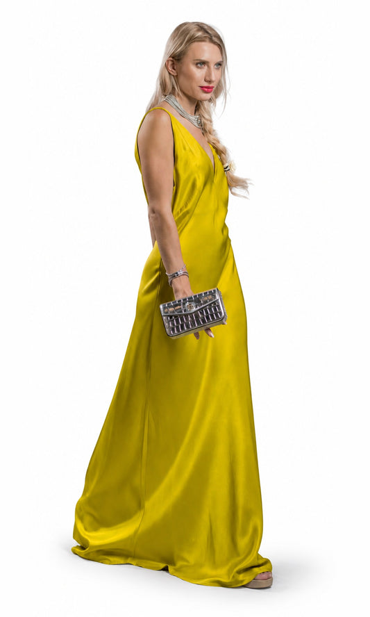 A blonde woman wearing a floor-length yellow satin gown with a deep V-neckline, holding a silver quilted clutch, photographed against a solid white background.