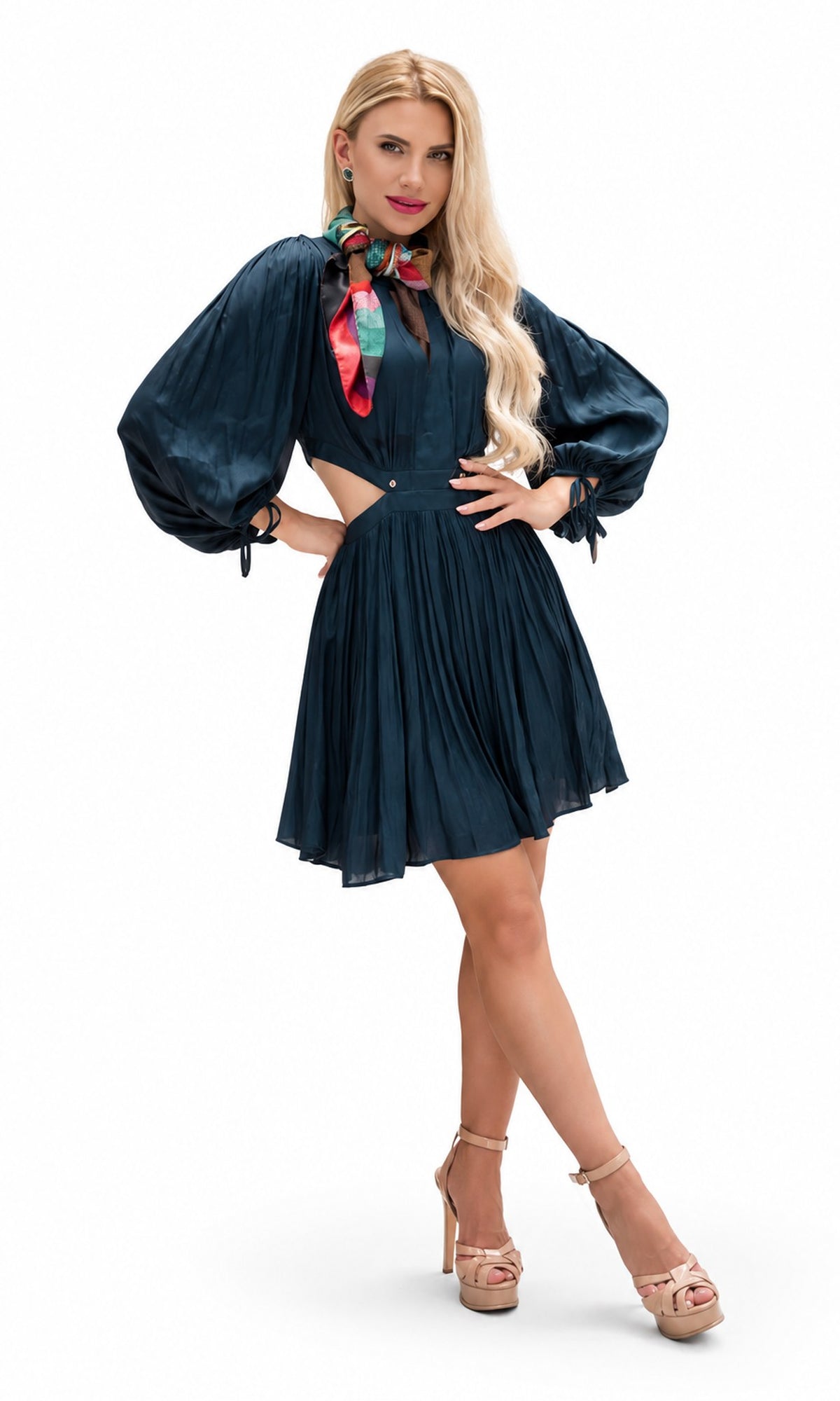 A woman modeling a dark teal blue pleated mini dress with voluminous puff sleeves, a high collar, and a cutout at the waist, styled with a colorful neck scarf and beige platform heels against a white studio background.