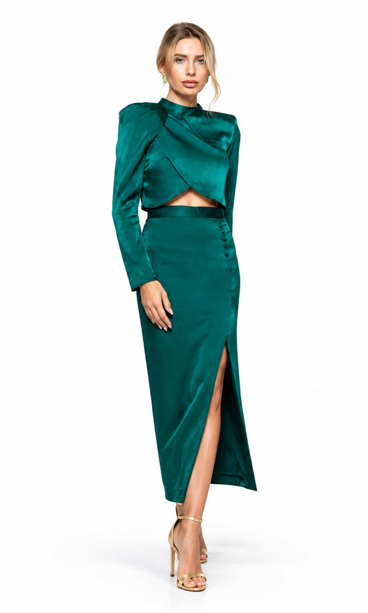 A woman modeling a dark emerald green satin two-piece outfit consisting of a long-sleeve cropped wrap-style top and a matching high-slit midi skirt, paired with nude high-heeled sandals against a plain white background.
