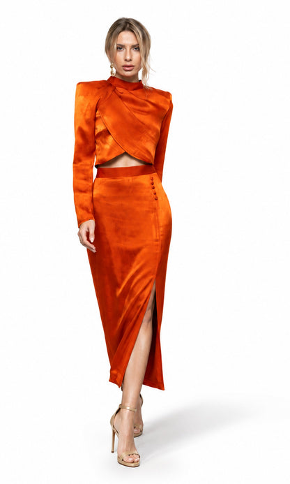 A woman modeling a bright orange satin two-piece set consisting of a long-sleeve wrap-style crop top and a matching high-slit midi skirt, paired with nude high-heeled sandals against a white studio background.